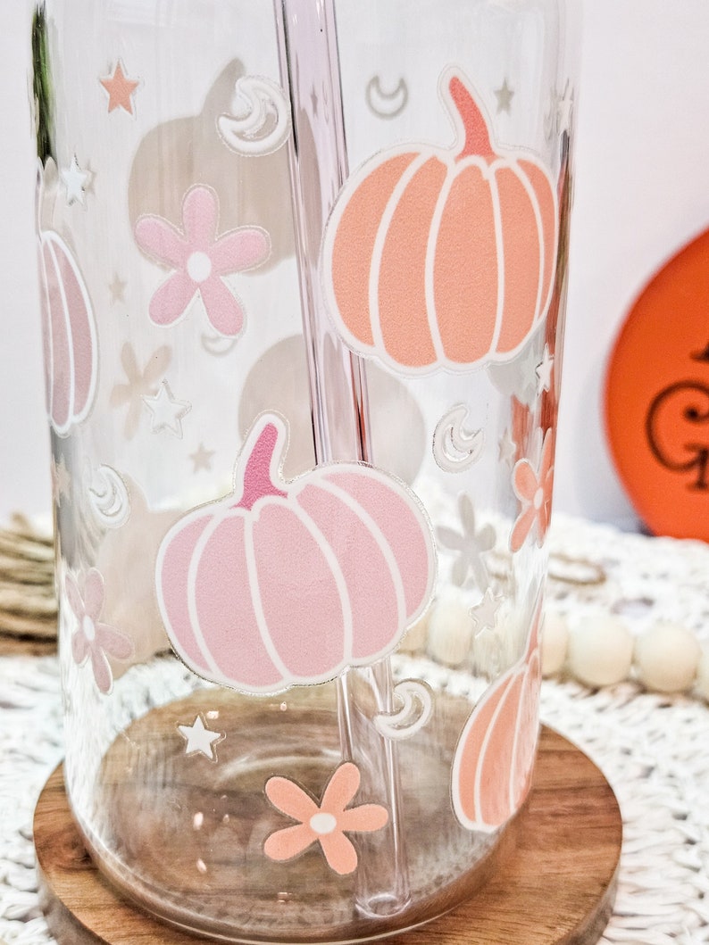 May include: A clear glass tumbler with a pink and orange pumpkin design. The tumbler has a straw and is sitting on a wooden coaster.