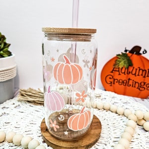 May include: A clear glass tumbler with a bamboo lid and a pink and orange pumpkin design. The tumbler has a straw and is sitting on a wooden coaster. The background is a white surface with a white and brown woven mat.
