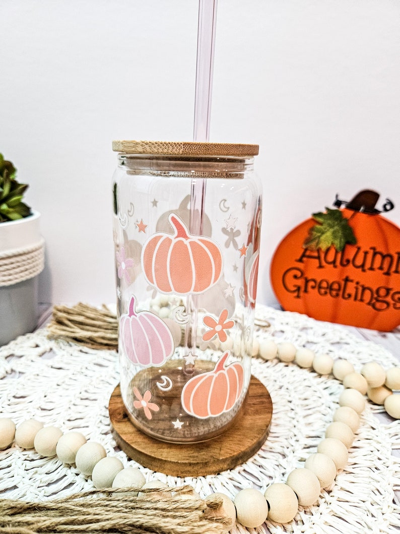 May include: A clear glass tumbler with a bamboo lid and a pink and orange pumpkin design. The tumbler has a straw and is sitting on a wooden coaster. The background is a white surface with a white and brown woven mat.