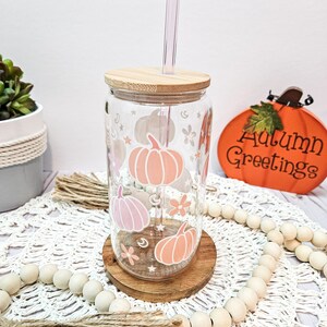 May include: A clear glass tumbler with a wooden lid and a straw. The tumbler has a design of pink and orange pumpkins, stars, and flowers. The tumbler is sitting on a wooden coaster and a white crocheted doily. The text "Autumn Greetings" is visible in the background.