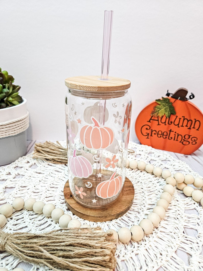 May include: A clear glass tumbler with a wooden lid and a straw. The tumbler has a design of pink and orange pumpkins, stars, and flowers. The tumbler is sitting on a wooden coaster and a white crocheted doily. The text "Autumn Greetings" is visible in the background.