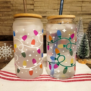 May include: Two frosted glass tumblers with bamboo lids and straws. Both tumblers feature a design of colorful Christmas lights on a white background. One tumbler has a clear design, while the other has a frosted design.