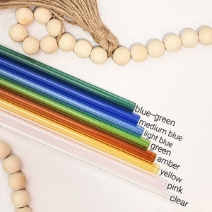 May include: A set of ten glass drinking straws in various colors, including blue-green, medium blue, light blue, green, amber, yellow, pink, and clear. The straws are arranged in a rainbow pattern and are displayed against a white background.