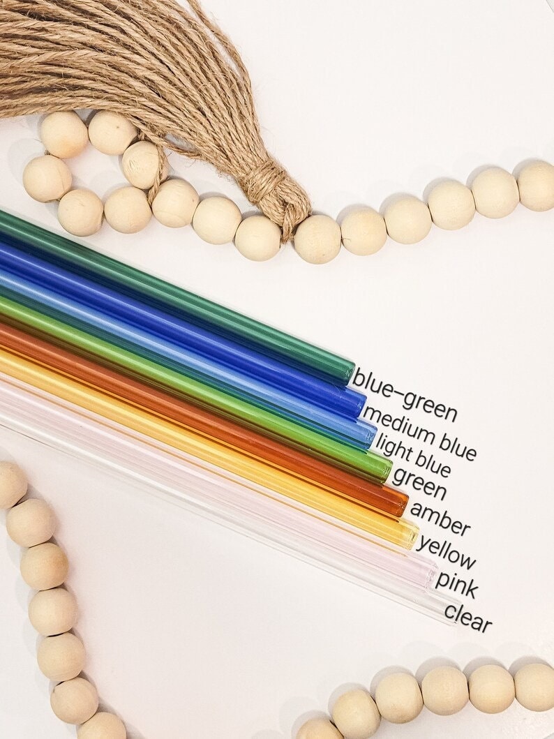 May include: A set of ten glass drinking straws in various colors, including blue-green, medium blue, light blue, green, amber, yellow, pink, and clear. The straws are arranged in a rainbow pattern and are displayed against a white background.