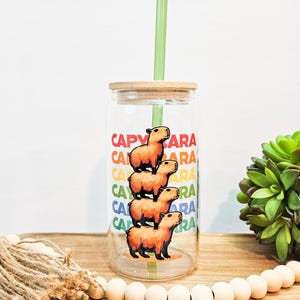 May include: A clear glass tumbler with a bamboo lid and a green straw. The tumbler has a stacked image of four capybaras and the word "CAPYBARA" repeated in rainbow colors.