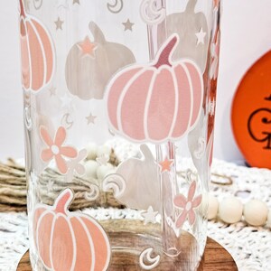 May include: A clear glass tumbler with a pink and white pumpkin design, stars, moons, and flowers. The tumbler has a straw in it and is sitting on a wooden coaster.