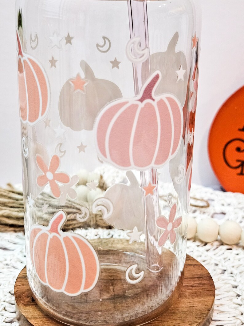 May include: A clear glass tumbler with a pink and white pumpkin design, stars, moons, and flowers. The tumbler has a straw in it and is sitting on a wooden coaster.
