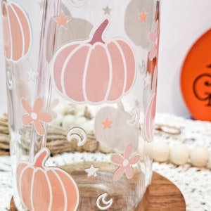May include: A clear glass tumbler with a pink and white pumpkin design, stars, moons, and flowers. The tumbler has a straw.