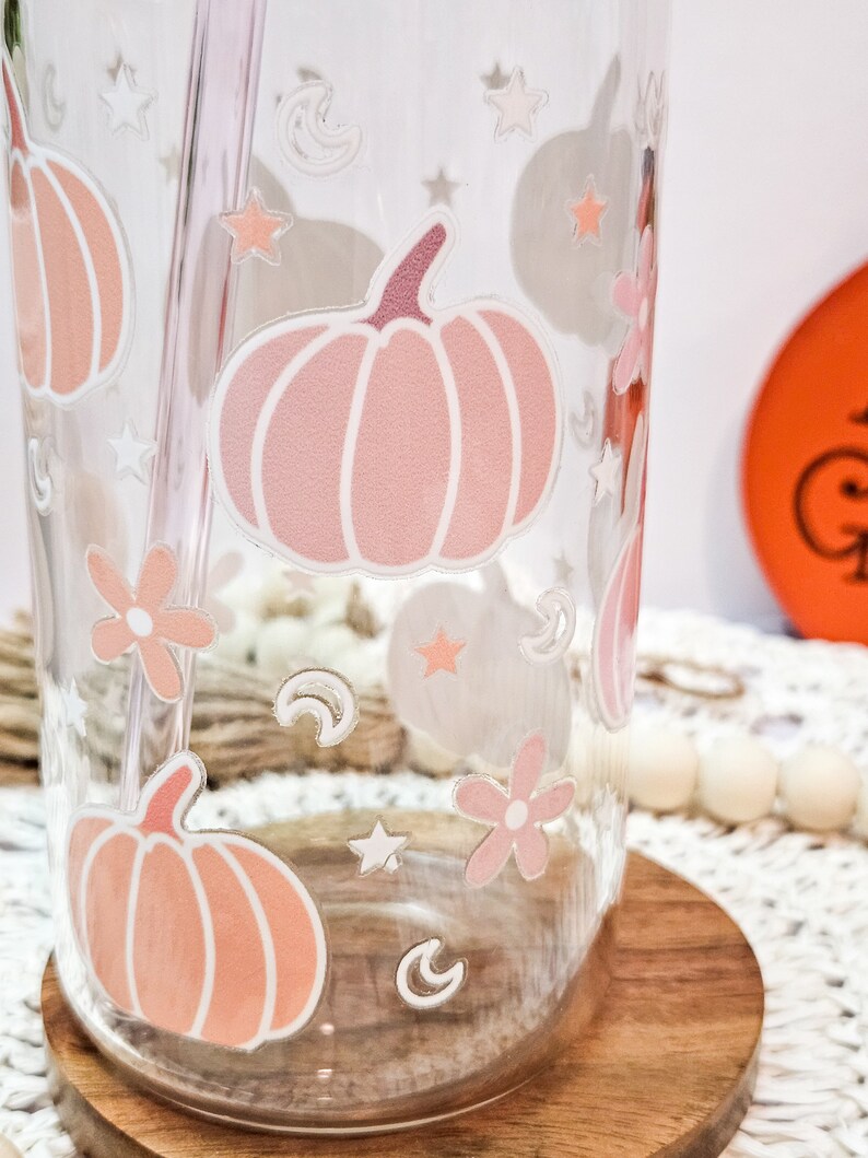 May include: A clear glass tumbler with a pink and white pumpkin design, stars, moons, and flowers. The tumbler has a straw.