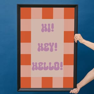Puede incluir: Obra de arte enmarcada con un diseño retro. La obra presenta un borde a cuadros naranja y rosa, con las palabras "HI!", "HEY!" y "HELLO!" en morado, sobre un fondo rosa claro. El marco es negro.