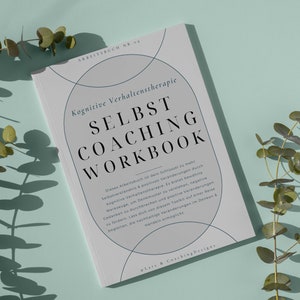 May include: A gray and white self-coaching workbook with the title "SELBST COACHING WORKBOOK" and the text "Kognitive Verhaltenstherapie" in black. The book is lying on a light blue surface with eucalyptus branches.