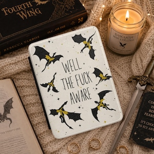 May include: A cream-colored tablet case with black and yellow dragon illustrations and the text "WELL THE FUCK AWARE." The image also features a book, a candle, a decorative sword, and rings, all arranged on a knitted surface.