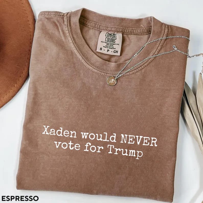 May include: A brown t-shirt with the text "Xaden would NEVER vote for Trump" printed in white on the front. The shirt is made of a soft, comfortable material and has a relaxed fit.