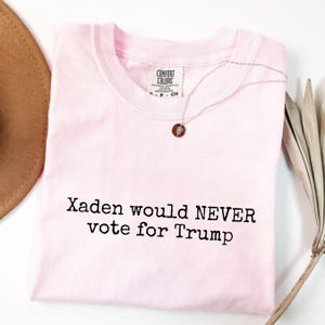 May include: Pink t-shirt with the text "Xaden would NEVER vote for Trump" printed in black.