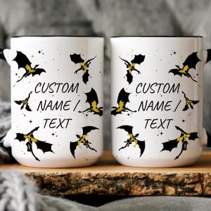 Puede incluir: Dos tazas de cerámica blanca con bordes y asas negros. Cada taza presenta un diseño de dragón negro y dorado con estrellas y el texto "CUSTOM NAME / TEXT".