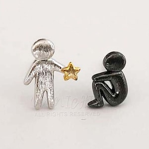 May include: A pair of silver and black earrings featuring a small person holding a gold star and another person sitting down.