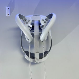 May include: A white VR headset with two gray controllers attached to the front. The controllers have buttons and a joystick. The headset is hanging from a wall mount.