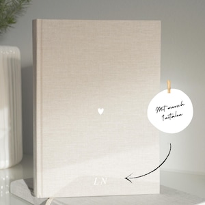 Könnte beinhalten: Ein beiges, mit Leinen bezogenes Buch mit einem kleinen weißen Herzen und den Initialen "LN" auf dem Einband. Ein rundes Etikett mit dem Text "Mit wunsch Initialen" hängt oben. Eine weiße Vase und ein kleiner Nadelbaumzweig sind im Hintergrund.