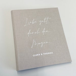 Könnte beinhalten: Ein hellgraues Hardcover-Buch mit dem Text "Liebe geht durch den Magen" in weißer Schrift. Die Namen "CLARA & THOMAS" sind darunter gedruckt. Das Buch hat einen strukturierten Einband.