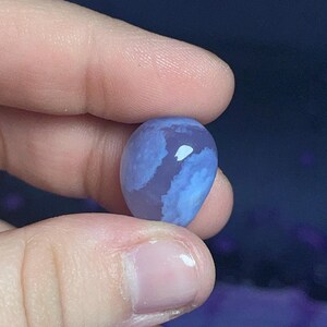 May include: A smooth, oval-shaped blue gemstone with white and lighter blue marbling. The stone is held between two fingers against a dark blue background. The gemstone's surface reflects light, highlighting its unique patterns.