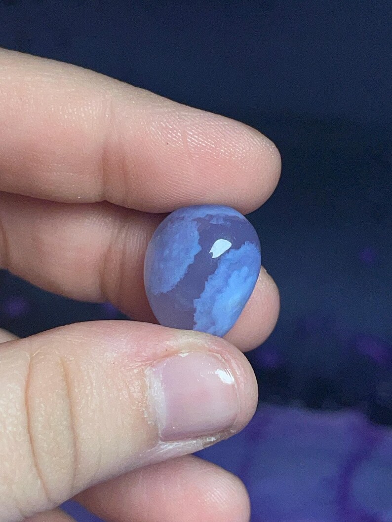 May include: A smooth, oval-shaped blue gemstone with white and lighter blue marbling. The stone is held between two fingers against a dark blue background. The gemstone's surface reflects light, highlighting its unique patterns.