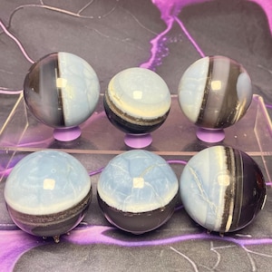 May include: Six polished gemstone spheres. The spheres are a mix of light blue, black, and white. Each sphere is mounted on a small, purple stand. The spheres have a marbled appearance and are arranged in two rows of three.
