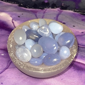 May include: A collection of smooth, polished blue chalcedony stones in varying shades of blue and gray, filling a small, frosted glass bowl. The bowl sits on a purple and white patterned fabric, creating a serene and natural aesthetic.