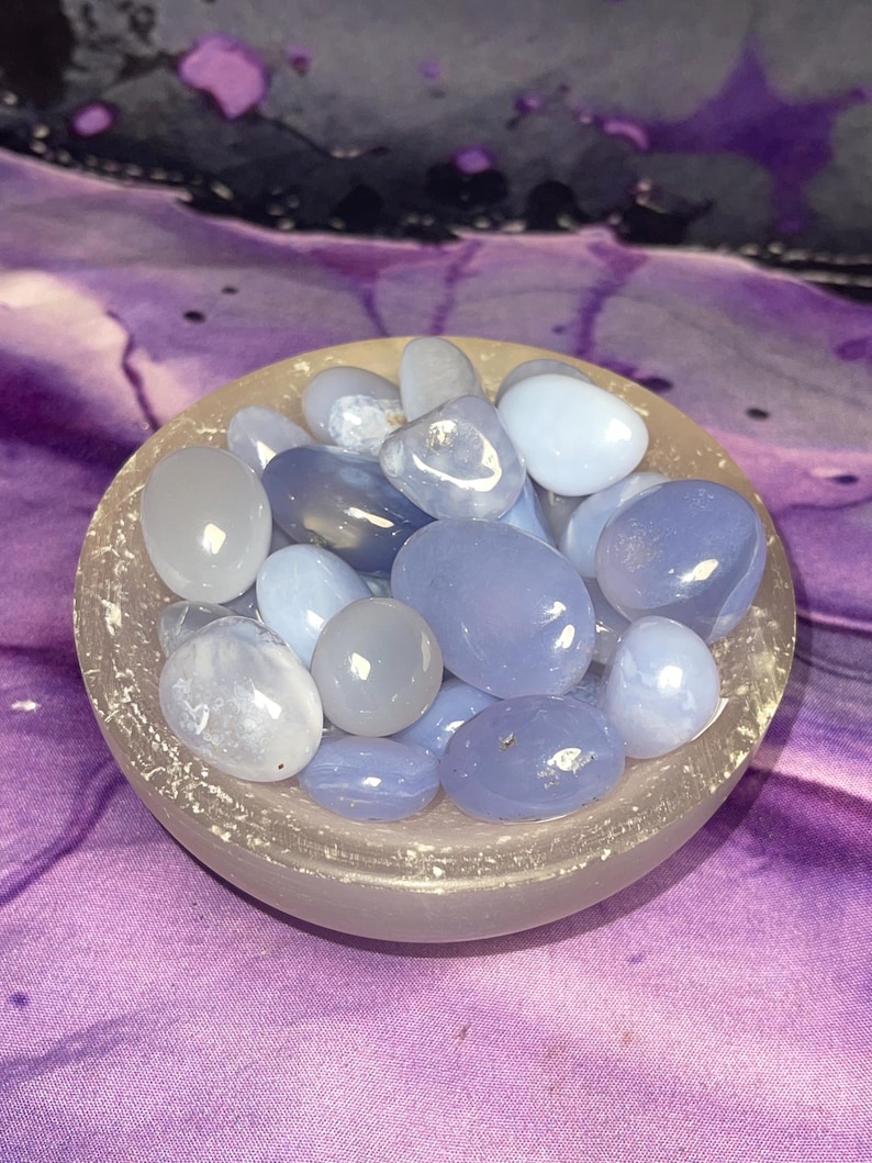 May include: A collection of smooth, polished blue chalcedony stones in varying shades of blue and gray, filling a small, frosted glass bowl. The bowl sits on a purple and white patterned fabric, creating a serene and natural aesthetic.