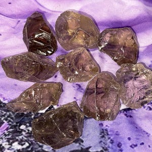 May include: A collection of rough-cut amethyst crystals in varying sizes and shapes. The crystals display shades of purple and brown, with a translucent quality. They are arranged on a purple fabric with a subtle pattern.