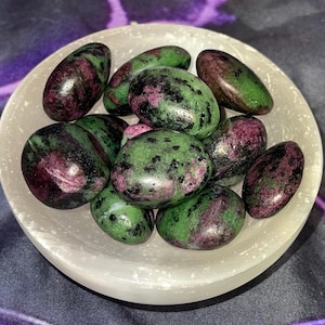 May include: A collection of polished ruby zoisite stones in a white, shallow bowl. The stones are oval-shaped, with a mix of green, dark pink, and black colors. The bowl sits on a dark, patterned surface.