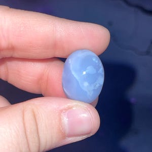 May include: A light blue, oval-shaped gemstone with white marbling. The stone is held between fingers against a dark blue background. The gemstone is smooth and polished, showcasing natural patterns.