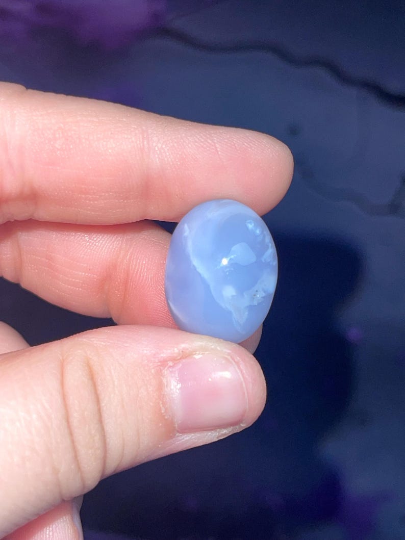 May include: A light blue, oval-shaped gemstone with white marbling. The stone is held between fingers against a dark blue background. The gemstone is smooth and polished, showcasing natural patterns.
