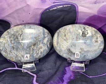 May include: Two oval-shaped, polished stones with a blue-grey and white marbled pattern. Each stone is displayed on a clear acrylic stand. The stones are labeled "A" and "B" with white text.