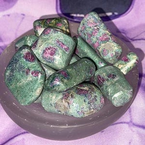 May include: A collection of green and pink tumbled stones with red ruby inclusions. The stones are in a gray bowl on a purple background.