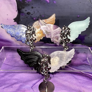 May include: A collection of gemstone angel wing pendants in various colors, including blue, black, white, and purple. Each pendant features a silver-toned figure and a circular base. The wings are made of different types of stone.