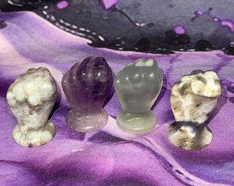 May include: Four carved stone figurines shaped like clenched fists standing in a row. The first is mottled white and gray, the second is translucent purple, the third is a pale translucent green, and the fourth is a marbled brown, white, and black.