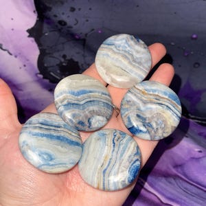 May include: Five round, polished stones with a swirling pattern of blue, white, and gray. The stones are held in a hand, showcasing their smooth, polished surfaces. The stones are a natural product.