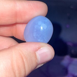 May include: A smooth, oval-shaped, light blue gemstone. The stone has a translucent quality with subtle white inclusions, and is held between fingers against a dark background. The gemstone is likely a polished cabochon.