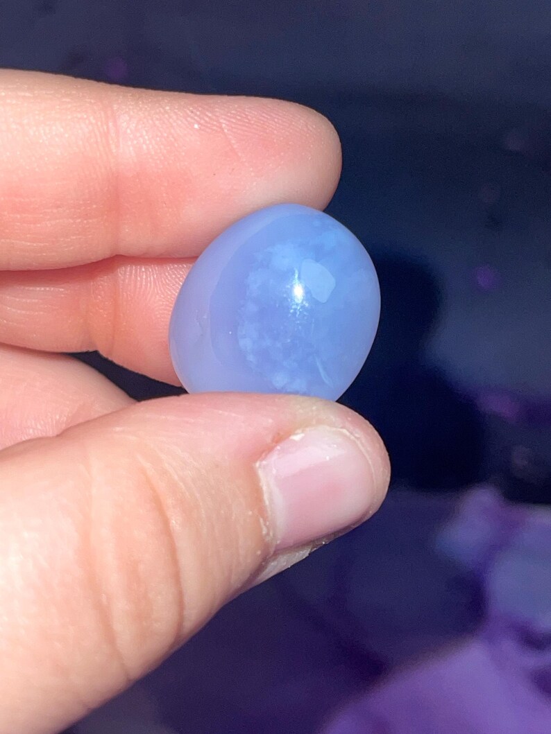 May include: A smooth, oval-shaped, light blue gemstone. The stone has a translucent quality with subtle white inclusions, and is held between fingers against a dark background. The gemstone is likely a polished cabochon.