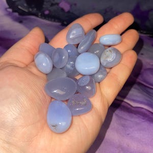 May include: A handful of smooth, polished blue chalcedony stones. The stones vary in shades of blue, from pale to a deeper periwinkle. Some stones have subtle white marbling. The stones are held in an open hand against a purple background.