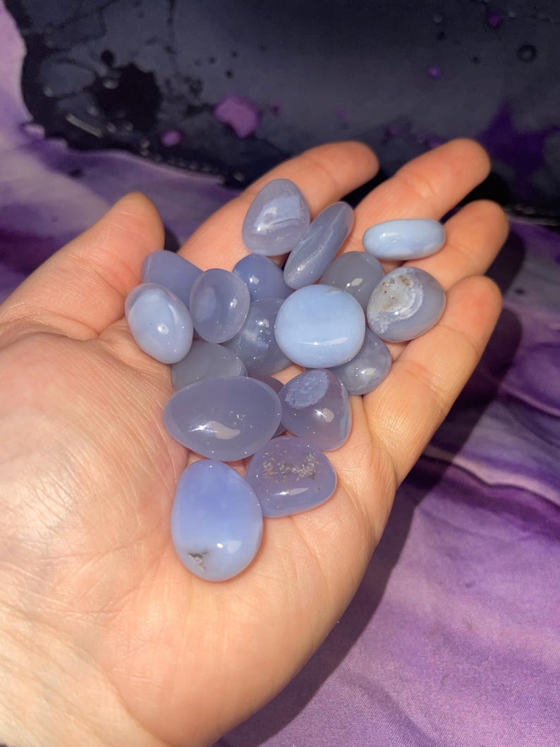 May include: A handful of smooth, polished blue chalcedony stones. The stones vary in shades of blue, from pale to a deeper periwinkle. Some stones have subtle white marbling. The stones are held in an open hand against a purple background.