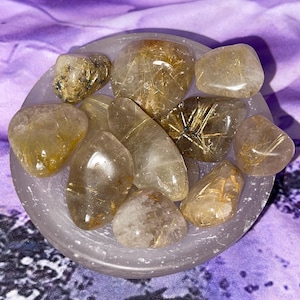 May include: A collection of polished, golden-brown rutilated quartz stones in a small, round, gray bowl. The stones have internal golden needle-like inclusions. The bowl sits on a purple patterned fabric.