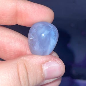 May include: A smooth, rounded, blue gemstone with white marbling is held between fingers. The stone has a polished surface and a soft, translucent appearance. The gemstone is a light periwinkle color.