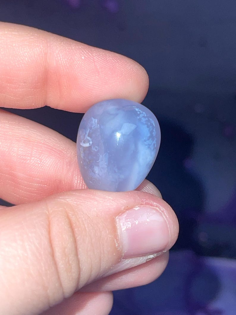 May include: A smooth, rounded, blue gemstone with white marbling is held between fingers. The stone has a polished surface and a soft, translucent appearance. The gemstone is a light periwinkle color.