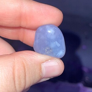 May include: A smooth, rounded, light blue gemstone is held between fingers. The stone has a polished appearance with subtle variations in color and texture. The gemstone is a soft, muted blue color.