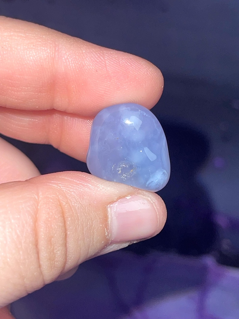 May include: A smooth, rounded, light blue gemstone is held between fingers. The stone has a polished appearance with subtle variations in color and texture. The gemstone is a soft, muted blue color.