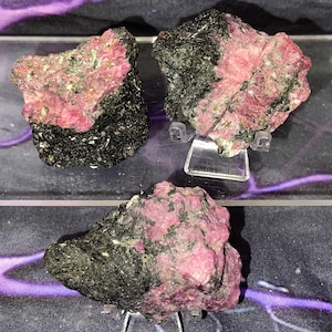 May include: Three raw mineral specimens. Each piece features a striking contrast of deep black and vibrant pink hues. The pink areas have a crystalline appearance, while the black sections appear dense. The minerals are displayed on clear stands.