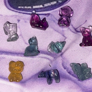 May include: A collection of small, carved gemstone animal figurines in various colors. The assortment includes a bear, cat, dog, and other animal shapes. The gemstones are shades of purple, blue, green, and yellow, set against a purple background.