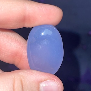 May include: A smooth, oval-shaped, light periwinkle-colored gemstone. The stone is held between two fingers, showcasing its polished surface and subtle color variations. The gemstone appears to be translucent, with a slight sheen.