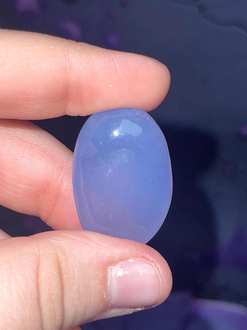 May include: A smooth, oval-shaped, light periwinkle-colored gemstone. The stone is held between two fingers, showcasing its polished surface and subtle color variations. The gemstone appears to be translucent, with a slight sheen.